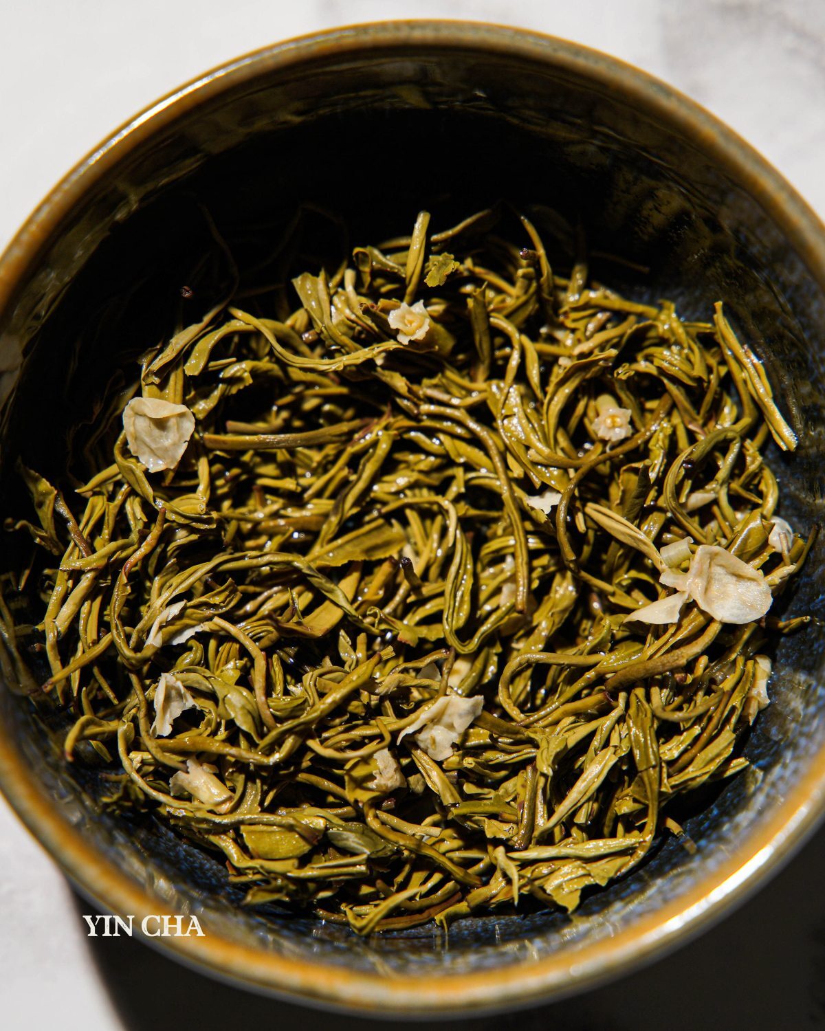 Loose jasmine green tea leaves with jasmine flowers in a dark ceramic bowl