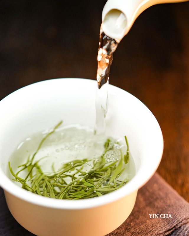 Hot water being poured over Steam Jade green tea leaves in a white teacup